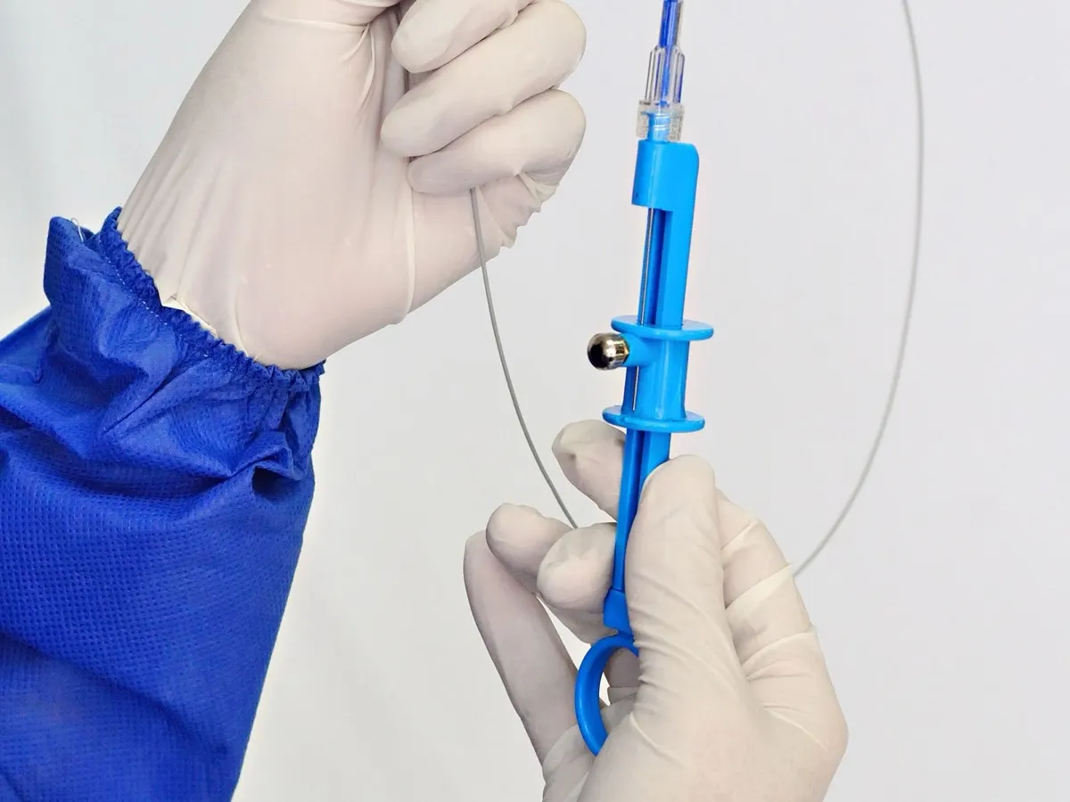Gloved hands in a blue gown holding a blue-handled catheter device with a thin guidewire looping beside it