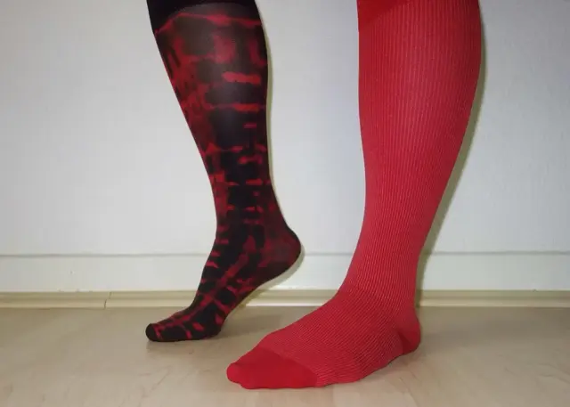 Lower legs and feet wearing knee-high compression socks, one red ribbed and one red and black patterned, against a white wall