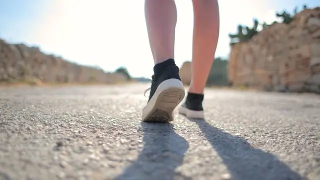 Low-angle view of a walker's bare calves and black sneakers stepping along a sunlit asphalt road