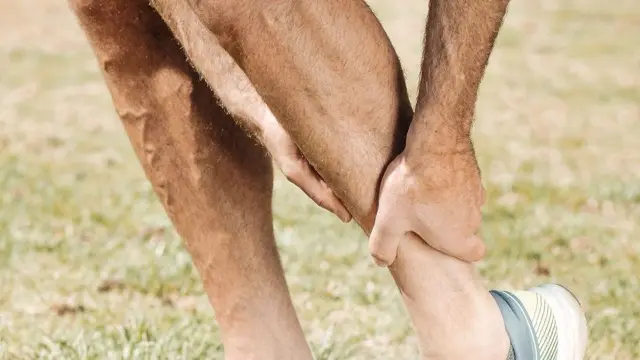 Close view of a man's bare calf gripped by both hands while he stands bent forward on grass in sneakers
