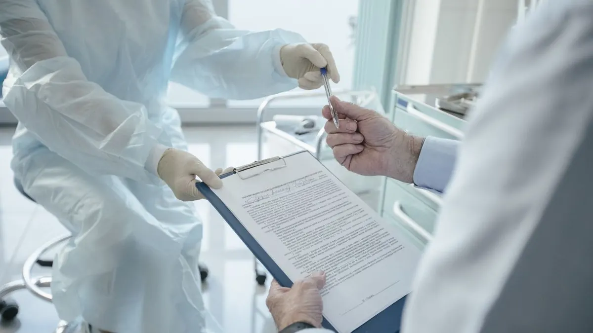Clinician in gloves and gown holding out a clipboard with a printed form and pen in a treatment room