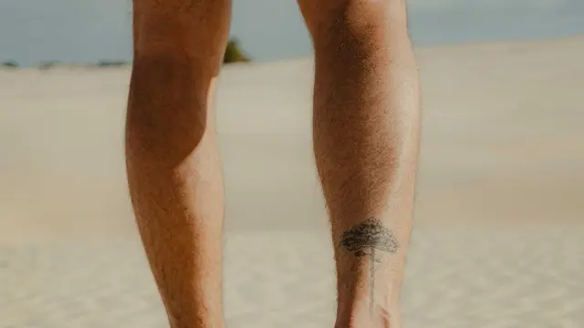 Back view of a man's bare calves with a small tree tattoo, walking barefoot on sand