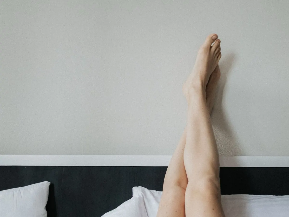Bare legs raised and crossed at the ankles against a plain wall above a bed with white pillows