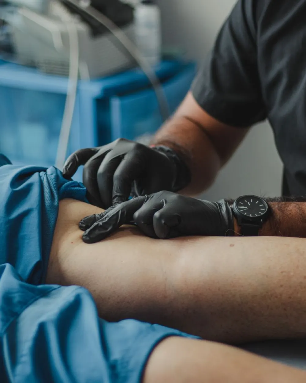 Clinician in scrubs and black gloves examining a patient's leg in a bright clinical room with blue equipment behind.